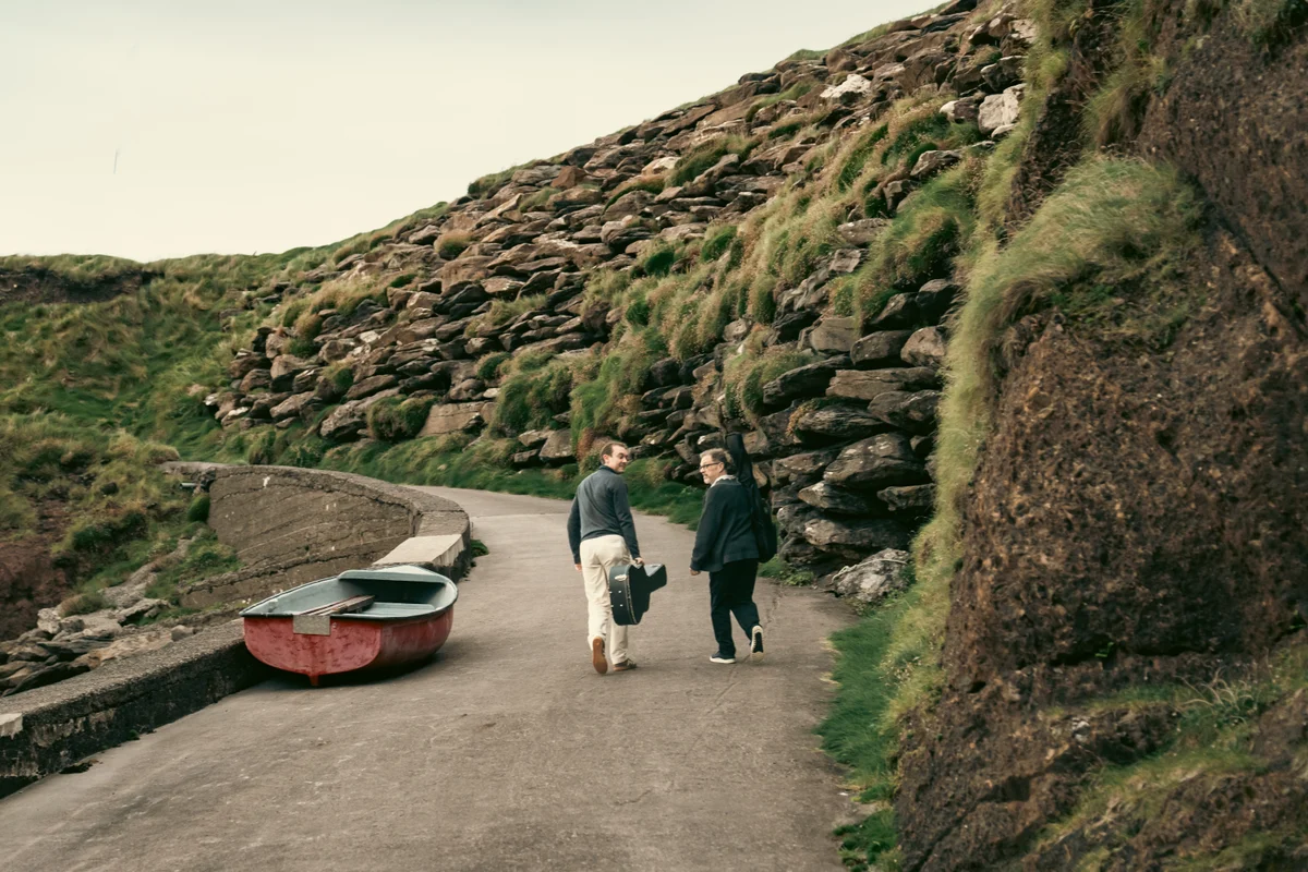 Dave and Tom carrying their instruments along a coastal path beside a stone wall